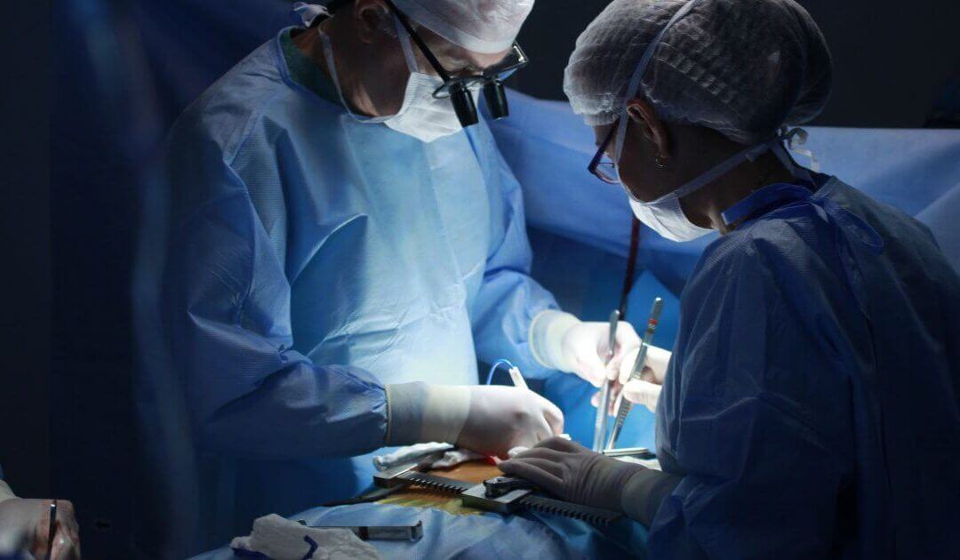 Two surgeons in blue scrubs perform a tummy tuck under bright lights, focusing intently on the patient. Surgical tools and equipment are visible in the foreground as they skillfully work to ensure a smooth post-surgery recovery.